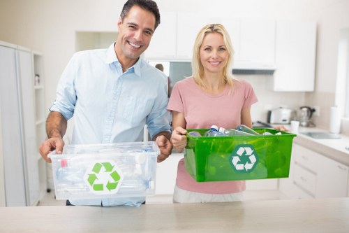 A man and a woman stand together in a bright, modern kitchen, each holding a transparent and a green plastic recycling bin with white recycling symbols on the sides. The man, positioned on the left, is wearing a light blue shirt and smiling, while the woman on the right, dressed in a pink top, also smiles and holds her bin securely. The kitchen background features white cabinets, a countertop with kitchen utensils, and a clean, organized environment. The scene emphasizes responsible waste separation and recycling practices, aligning with rubbish removal services provided by House Clearance Chislehurst in the Chislehurst area. The overall setting suggests a focus on environmentally conscious waste management in a domestic context, with good lighting and a neutral, inviting atmosphere.