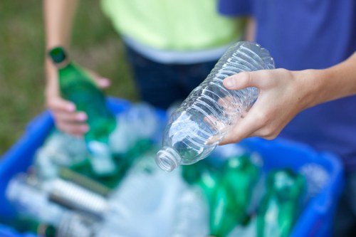 Close-up of a person's hand holding a clear plastic bottle, part of a collection of recyclable bottles, with a blue recycling bin filled with green and transparent plastic bottles and other waste materials visible in the background. The scene appears to be outdoors on a paved or grassy surface, with another individual partially visible wearing a yellow shirt, indicating a rubbish collection or recycling activity in a residential area near Chislehurst. The image emphasizes waste separation and recycling, reflecting services provided by House Clearance Chislehurst specializing in rubbish removal and clearance solutions in the local area near postcode BR7, with a focus on sustainable disposal practices.